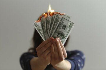 A woman holds burning hundred dollar bills in front of her face