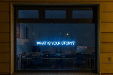 a neon sign reading "what is your story?" hangs in a darkened window